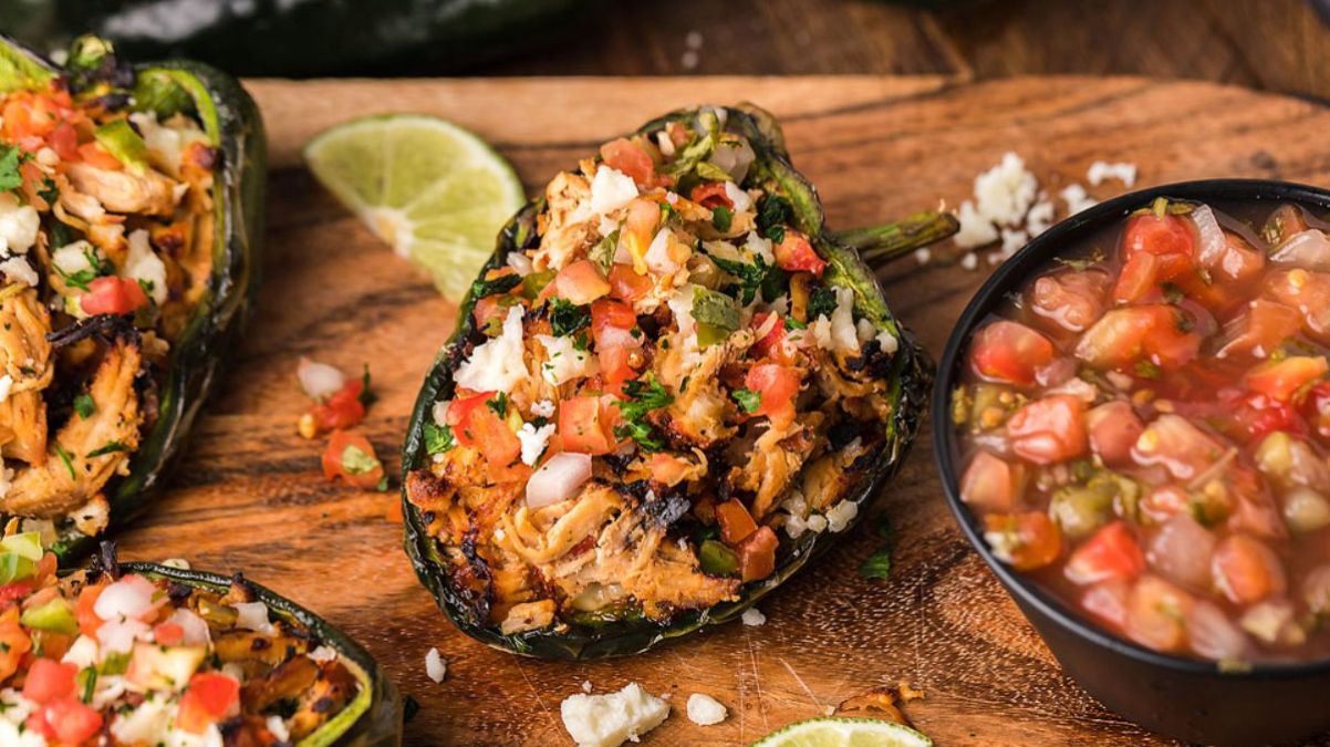 Halved poblano peppers stuffed with seasoned chicken and vegetables, garnished with cheese and herbs, served on a wooden board with lime slices and a bowl of salsa.