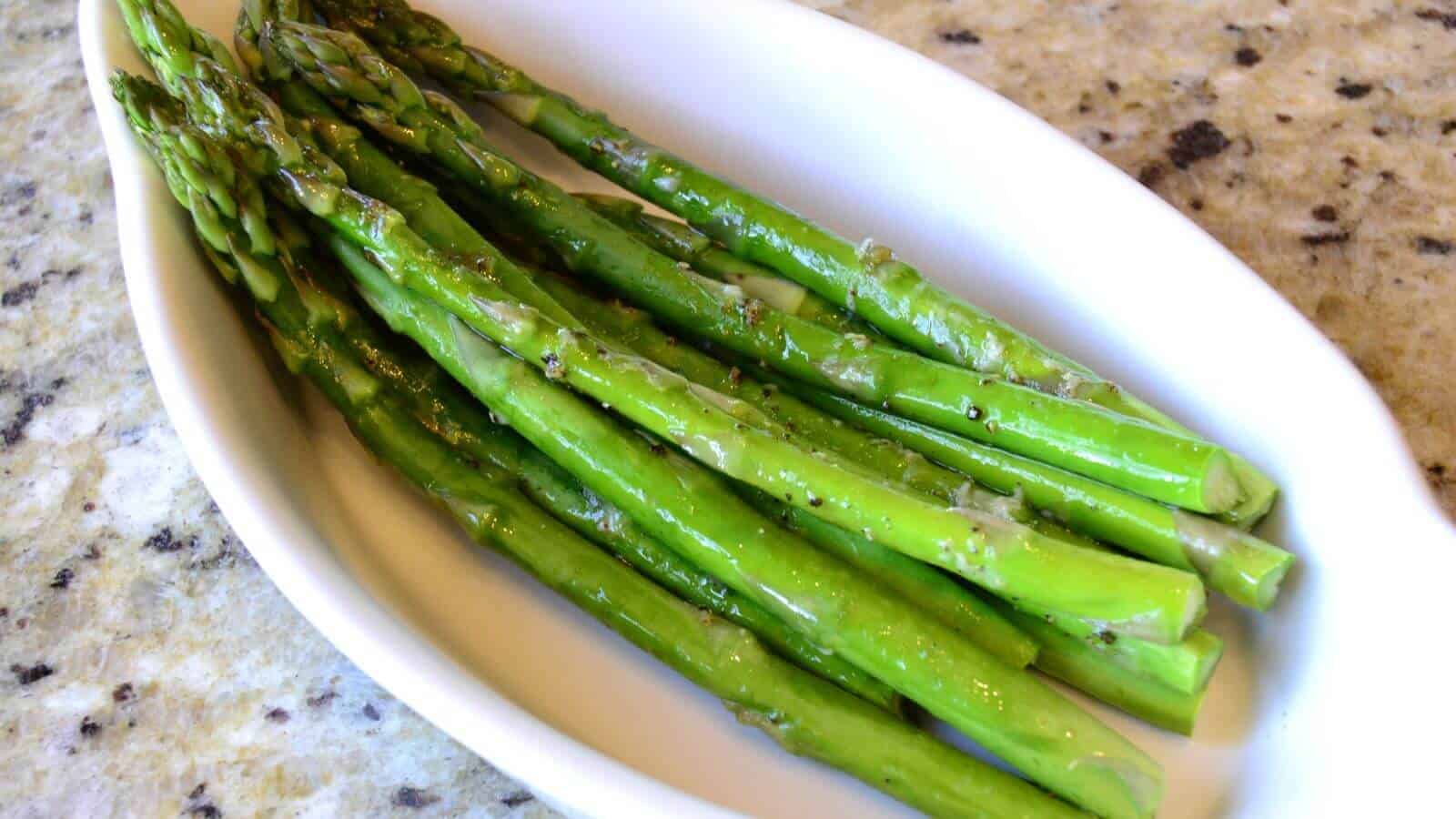 A white dish with several cooked asparagus spears seasoned with herbs, placed on a granite countertop.