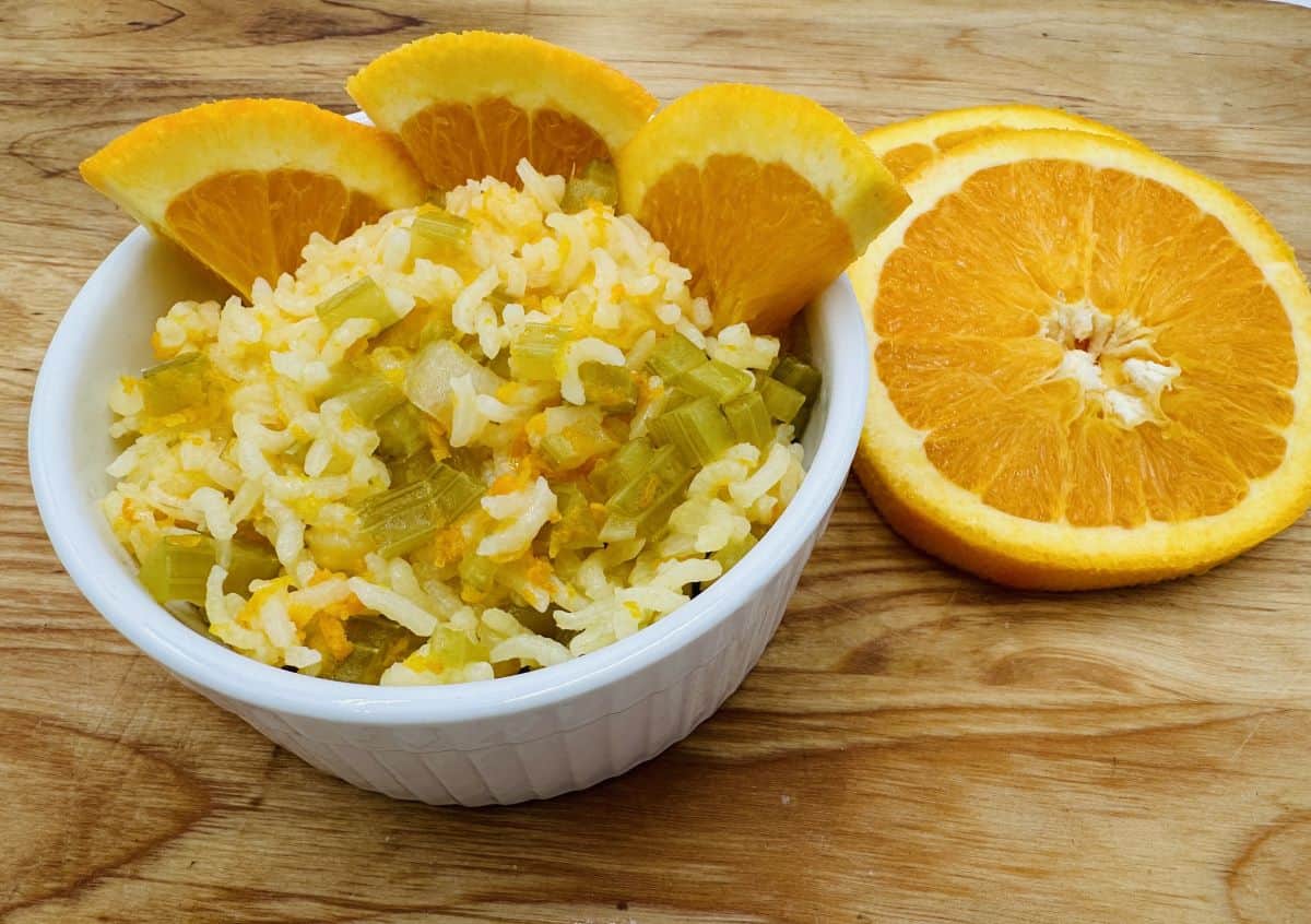 A white bowl of rice mixed with vegetables, garnished with orange slices, sits on a wooden surface next to additional orange slices.