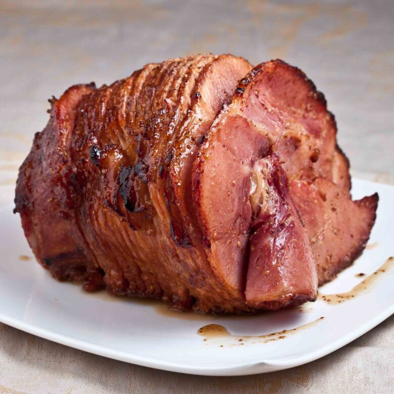 A sliced glazed ham sits on a white plate, with visible caramelized edges and juices on the surface.