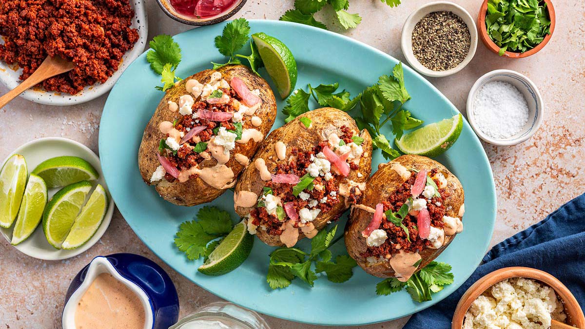 Three baked potatoes on a blue platter, topped with crumbled meat, cheese, sauce, herbs, and bacon, garnished with lime wedges and cilantro. Various toppings in bowls surround the platter.