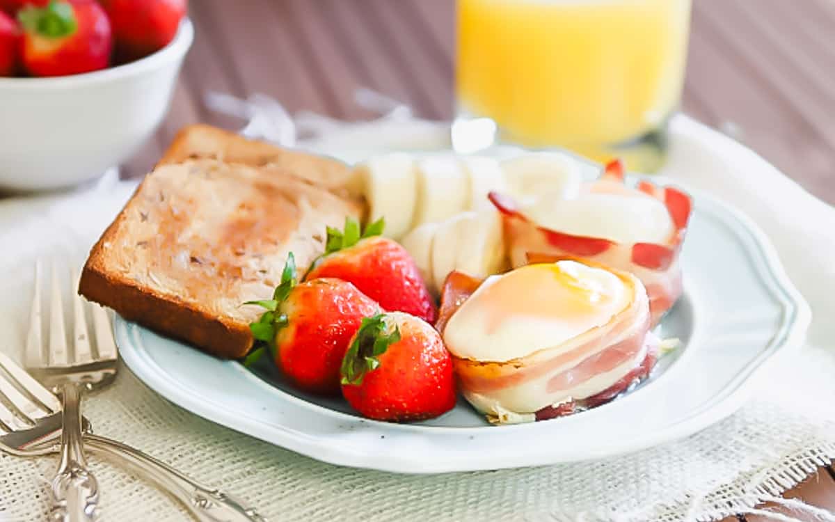A breakfast plate with toast, fresh strawberries, sliced pears, a bacon-wrapped egg, and a glass of orange juice in the background.