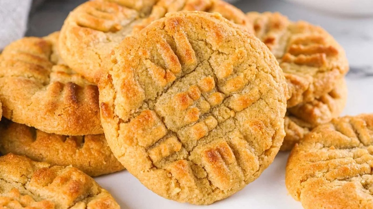 A close-up of several golden-brown peanut butter cookies with a crisscross fork pattern on top, stacked on a white surface.