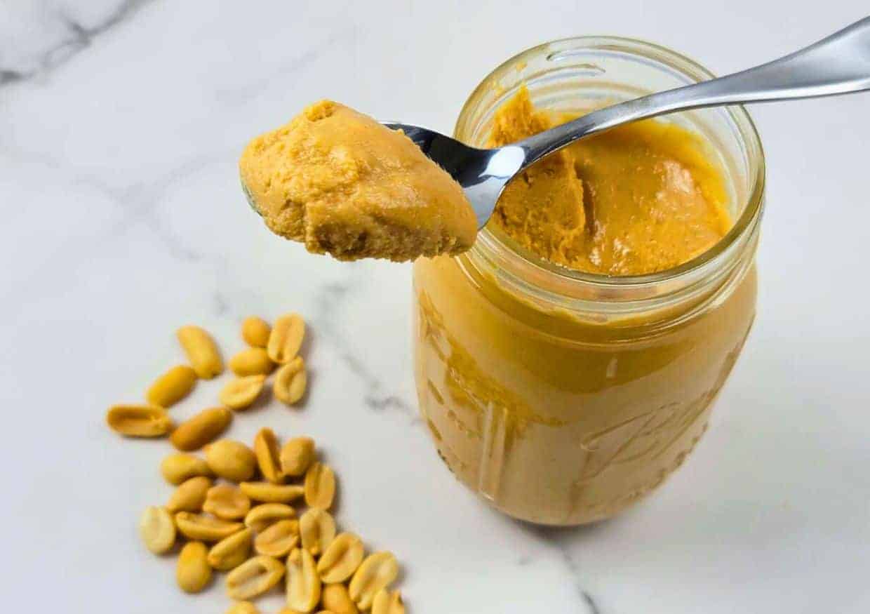 An open jar of peanut butter with a spoonful above it, next to a small pile of shelled peanuts on a white surface.