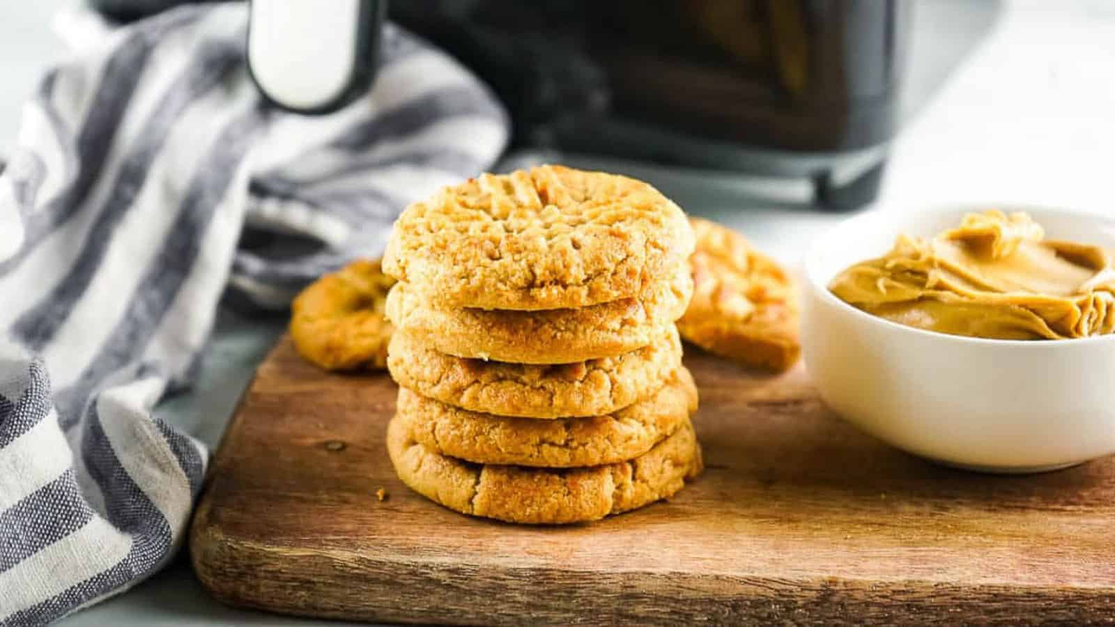 A stack of peanut butter cookies sits on a wooden board next to a bowl of peanut butter and a striped cloth.