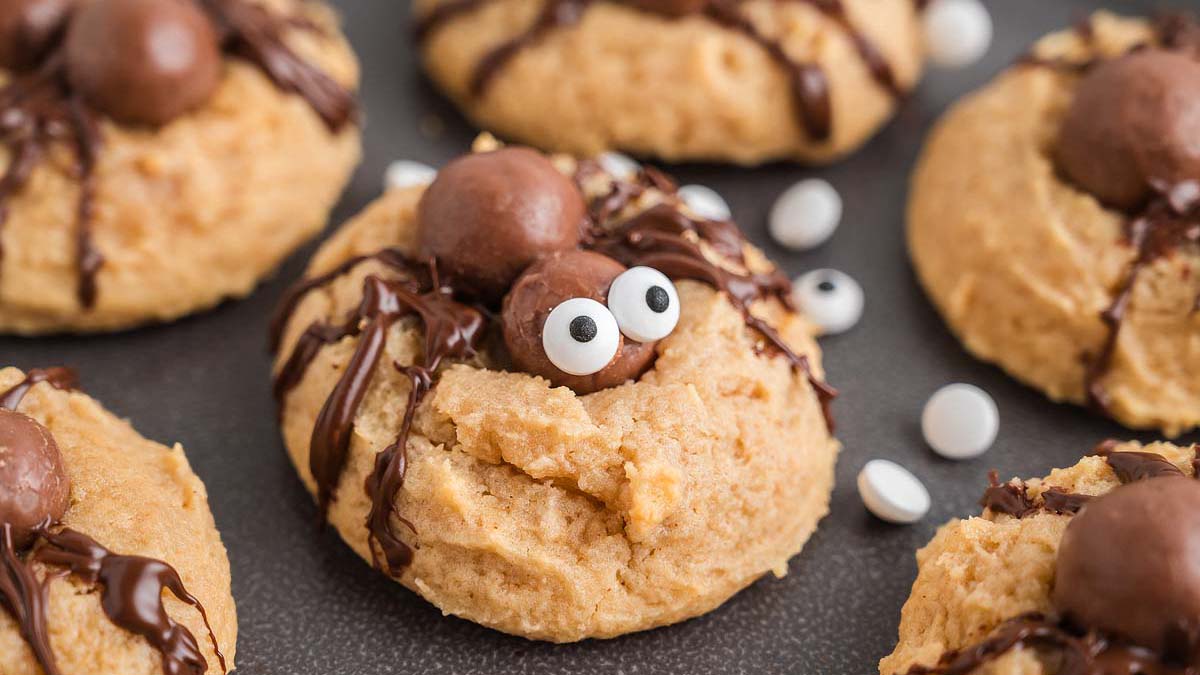 A close-up of peanut butter cookies decorated with chocolate, candy eyes, and chocolate balls to look like spiders on a dark surface.