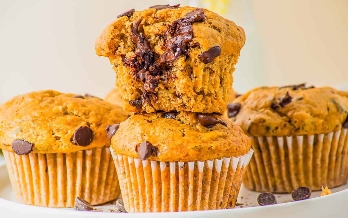 Three chocolate chip muffins are stacked on a plate, with the top muffin missing a bite, revealing its moist interior and melted chocolate chips.