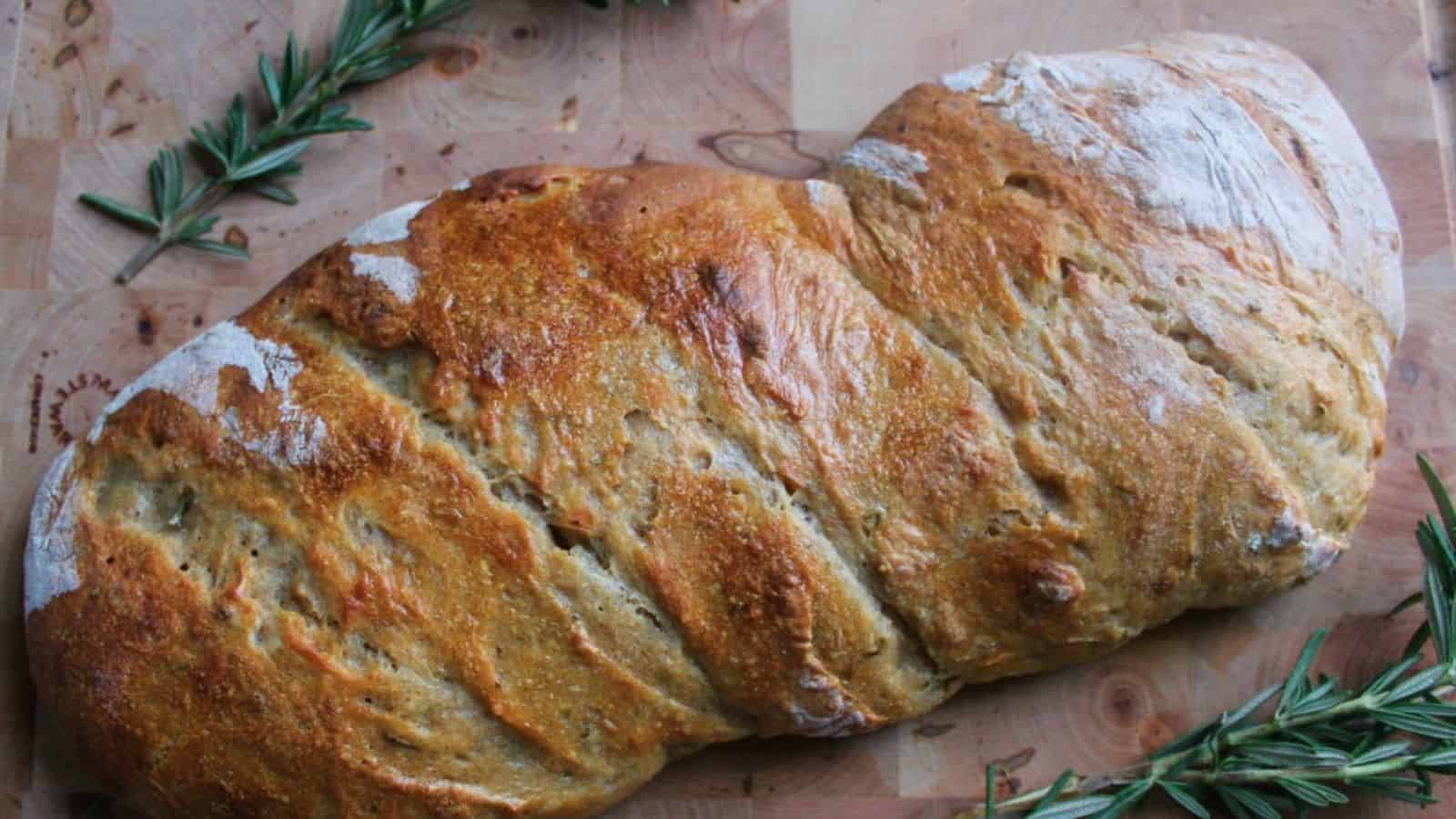 A rustic, golden-brown loaf of bread with a twisted shape sits on a wooden board, surrounded by sprigs of fresh rosemary.