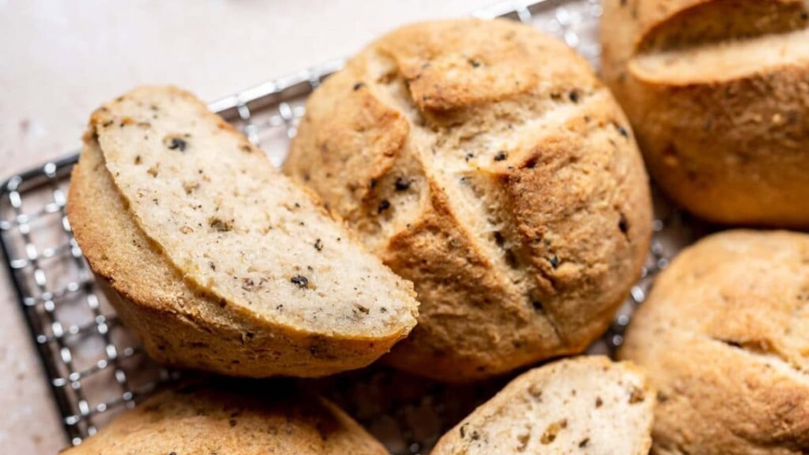 Round loaves of bread with a golden crust and visible herbs or seeds rest on a cooling rack; one loaf is sliced to show the inside texture.