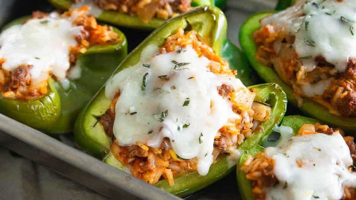 Green bell peppers halved and filled with a rice and meat mixture, topped with melted white cheese and herbs, arranged in a baking tray.