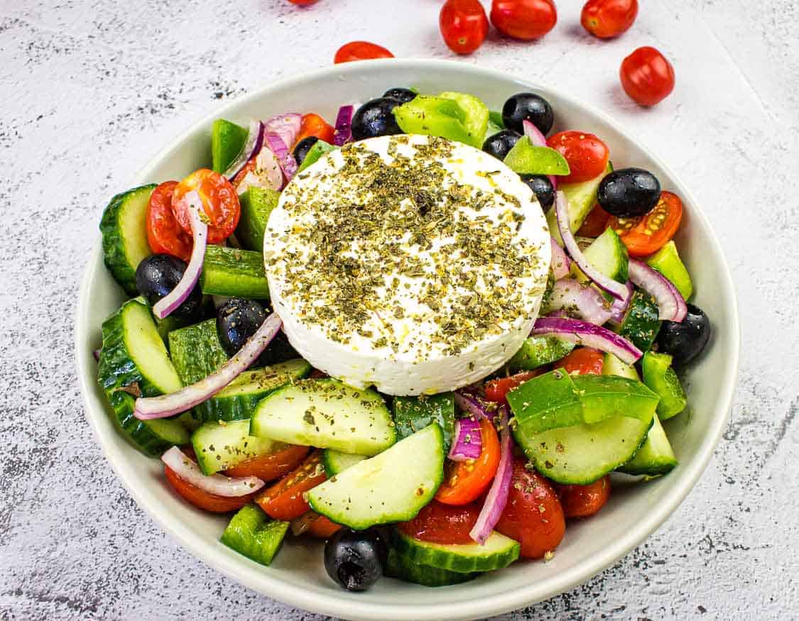 A bowl of Greek salad with cucumber, tomato, red onion, black olives, green pepper, and a round slice of feta cheese topped with herbs.
