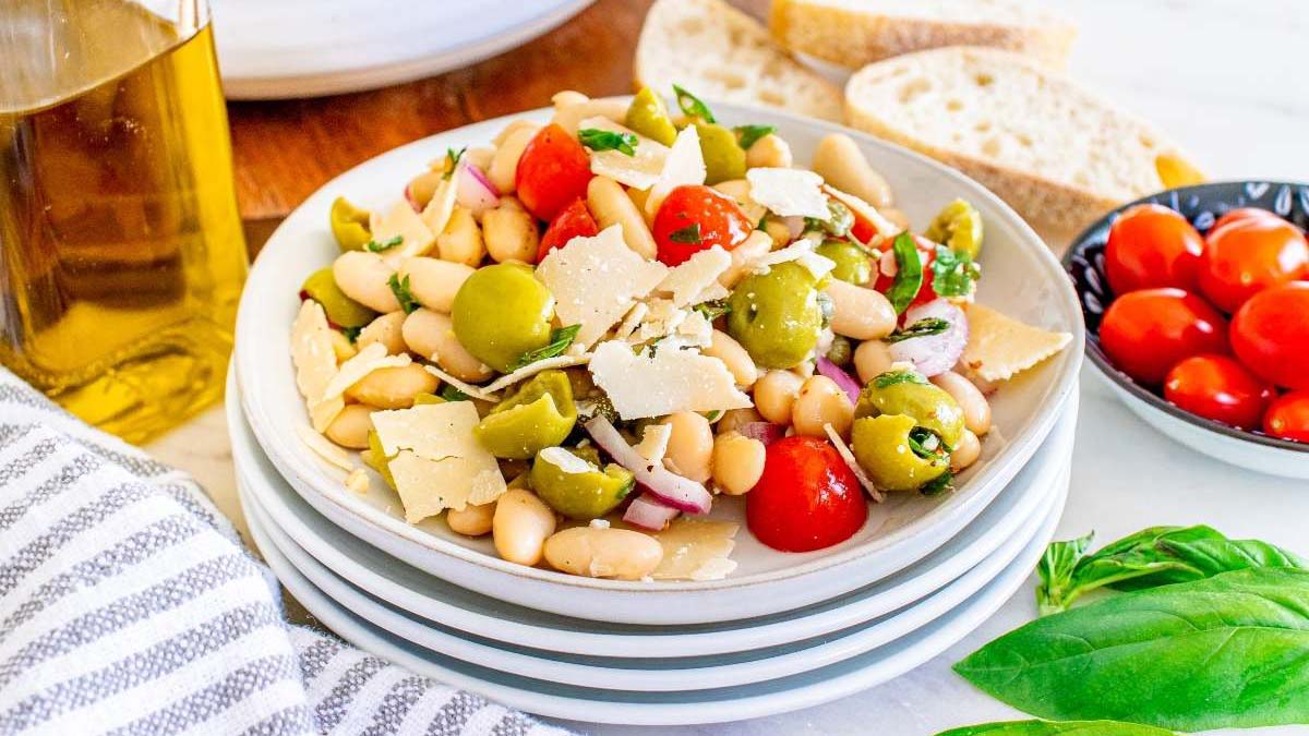 A plate of bean salad with cherry tomatoes, green olives, red onion, parsley, and parmesan flakes, served with slices of bread and a bowl of extra tomatoes in the background.