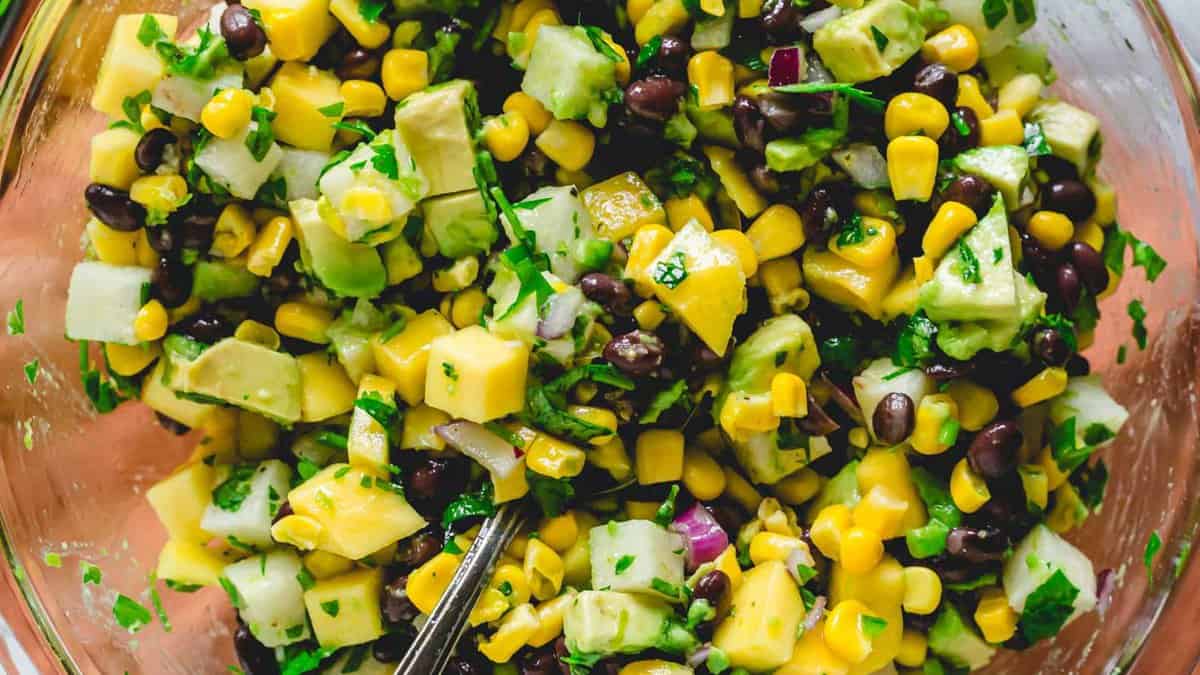 A bowl of salad containing avocado, corn, black beans, diced mango, red onion, cilantro, and cucumber, with a spoon visible.