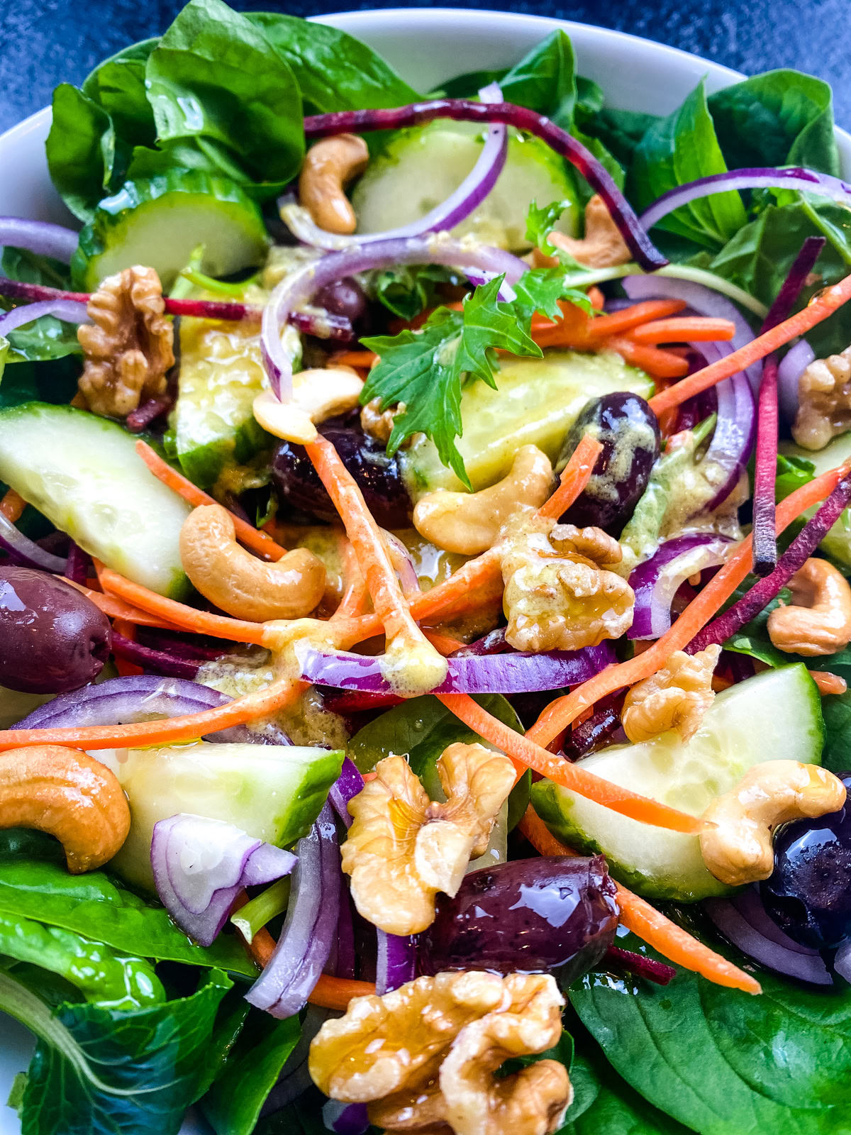 A salad with spinach, sliced cucumbers, shredded carrots, red onion, olives, walnuts, cashews, and parsley, topped with a light dressing.