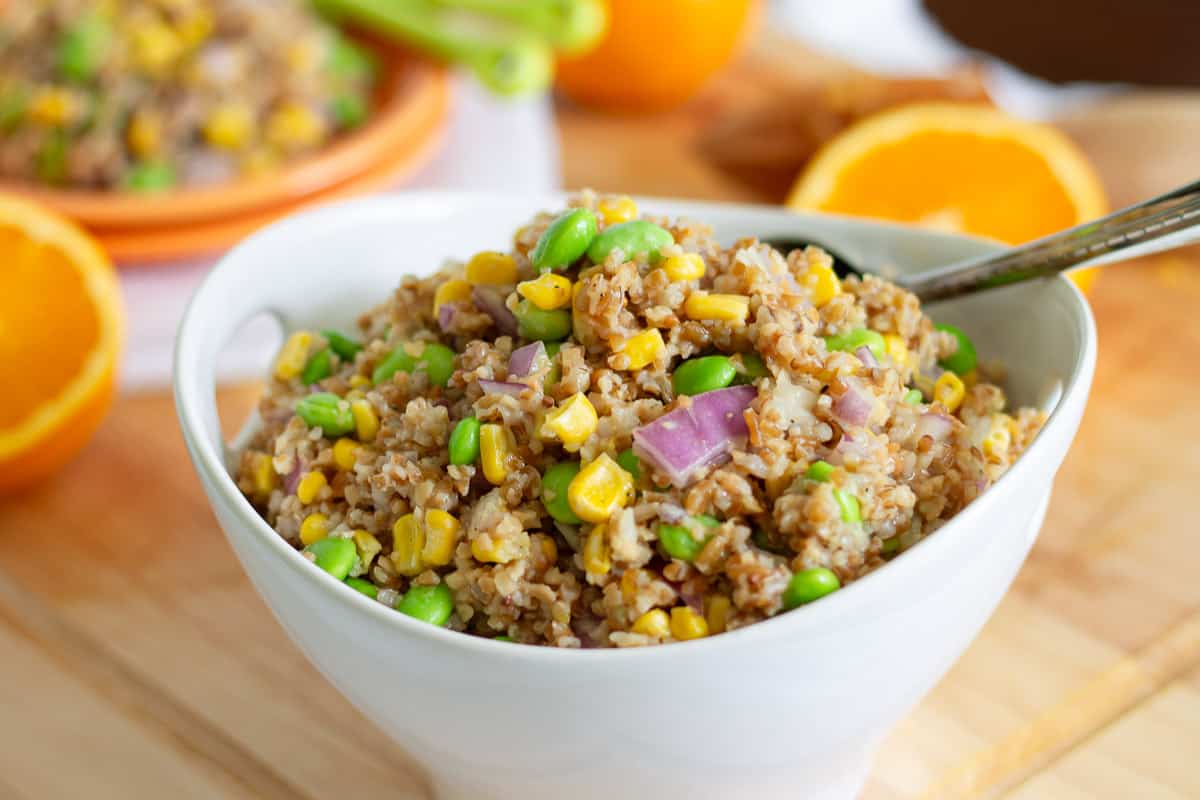 A white bowl filled with a mixture of brown rice, corn, edamame, and chopped red onions, with a spoon inside. Orange slices are visible in the background.