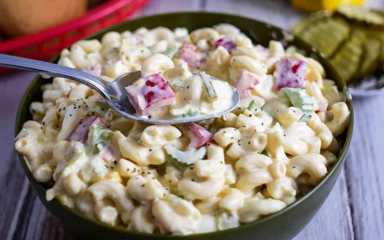 A bowl of creamy macaroni salad with chopped celery and red bell peppers, with a spoon holding a serving above the bowl.