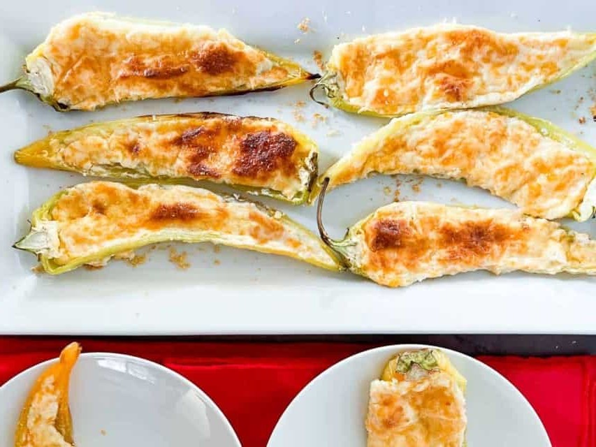 Six baked, cheese-stuffed peppers are arranged on a white tray, with two more on small plates, all set on a red tablecloth.