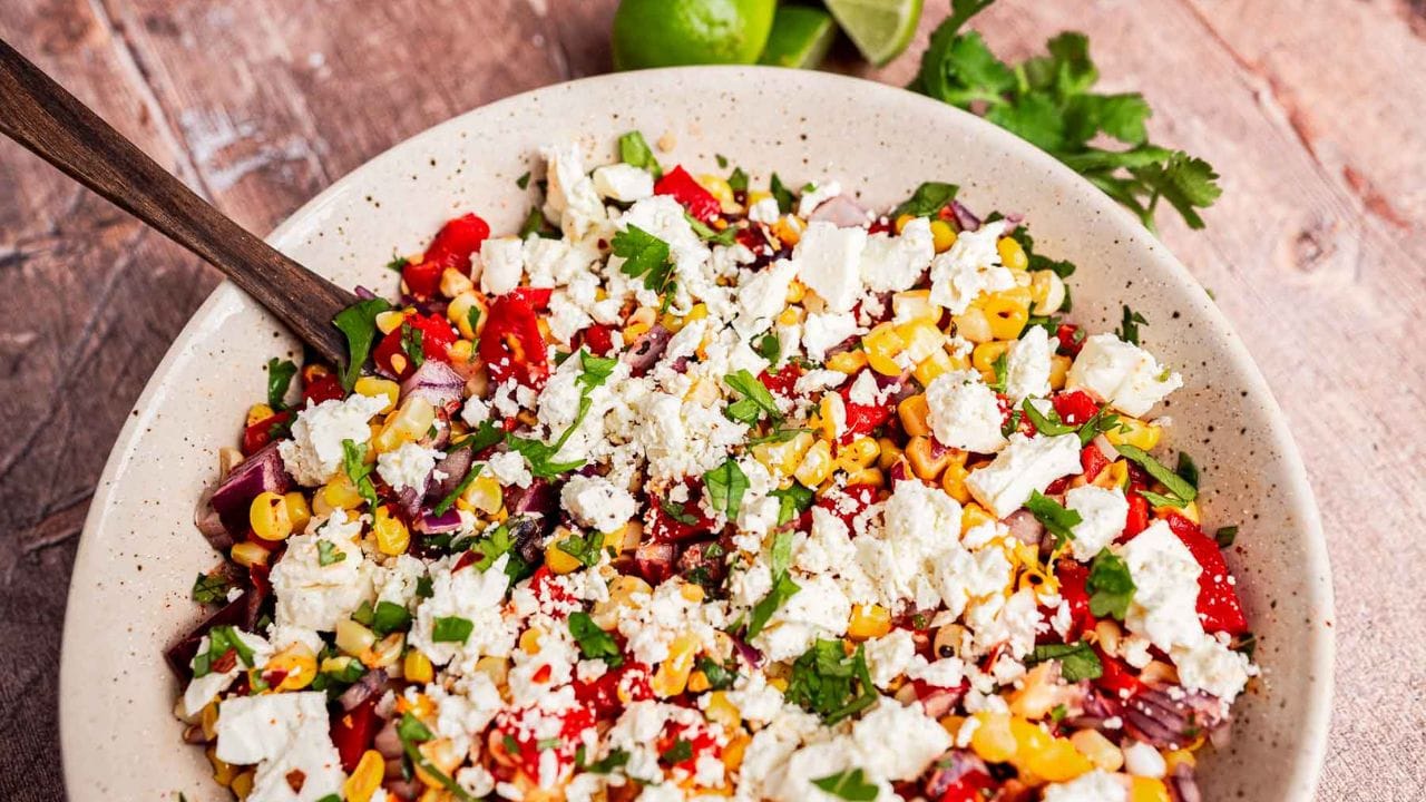 A bowl of salad with corn, diced red onion, red bell pepper, fresh cilantro, and crumbled feta cheese, served with a wooden spoon on a rustic wooden table.