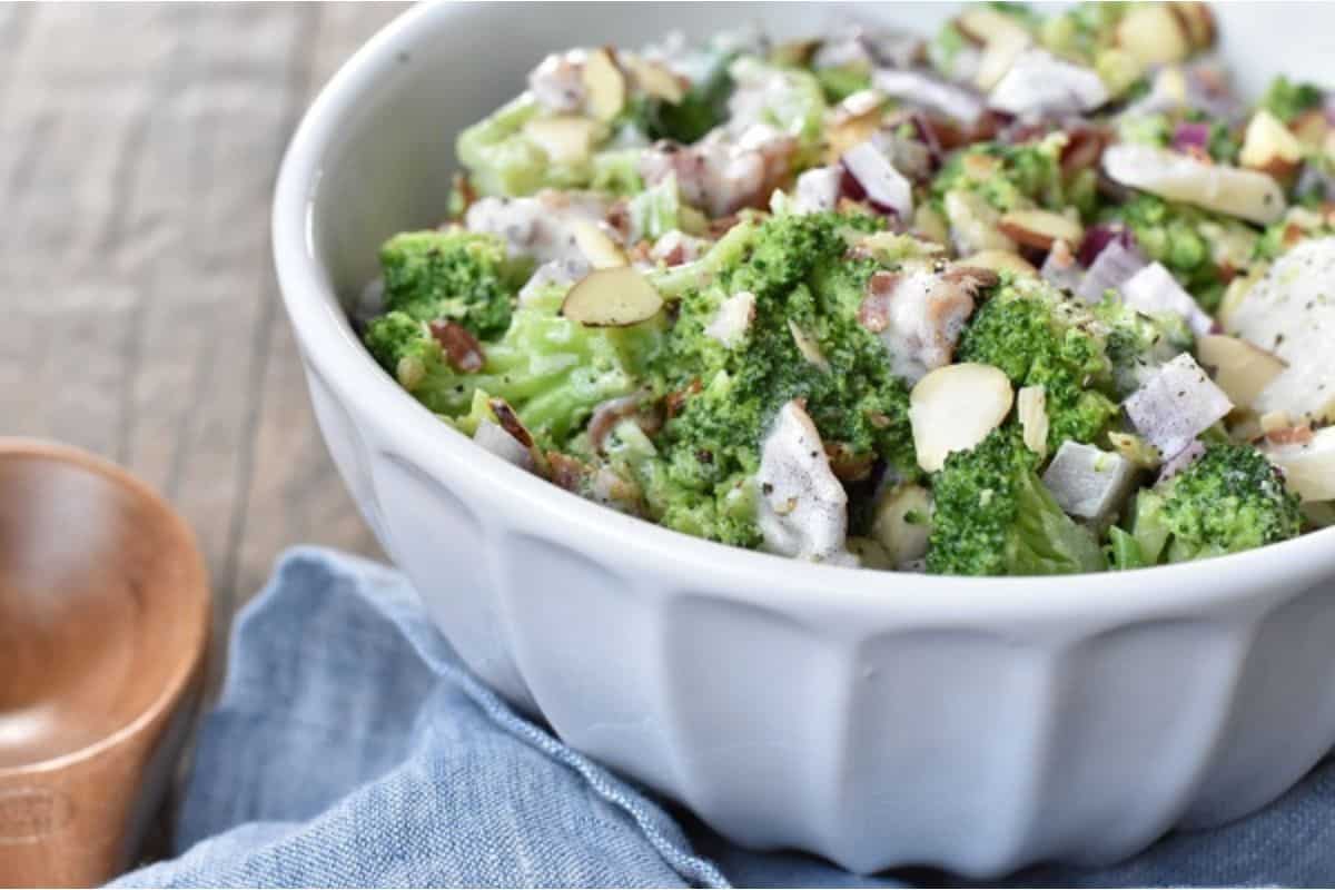 A white bowl filled with a broccoli salad topped with sliced almonds, red onions, and a creamy dressing sits on a blue cloth.