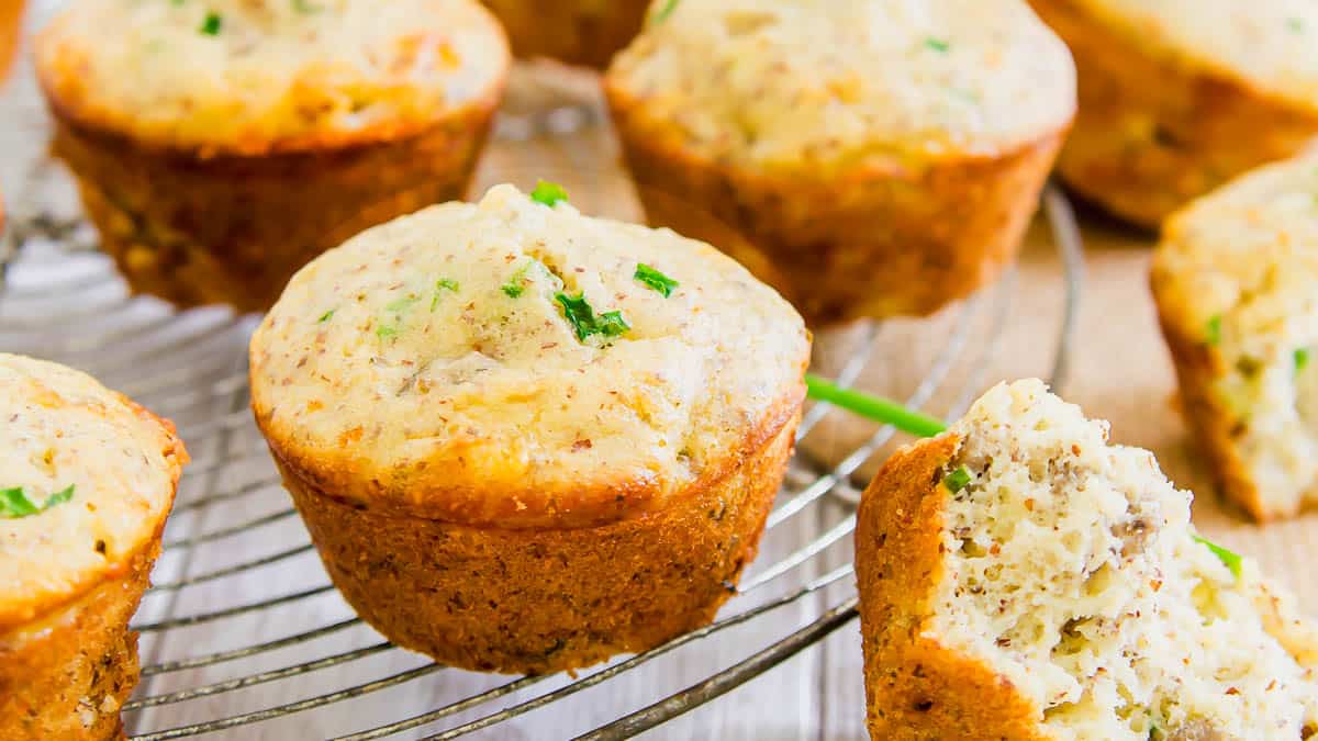 Several savory muffins with herbs are cooling on a wire rack, with one muffin cut open to show its moist interior and visible herbs.