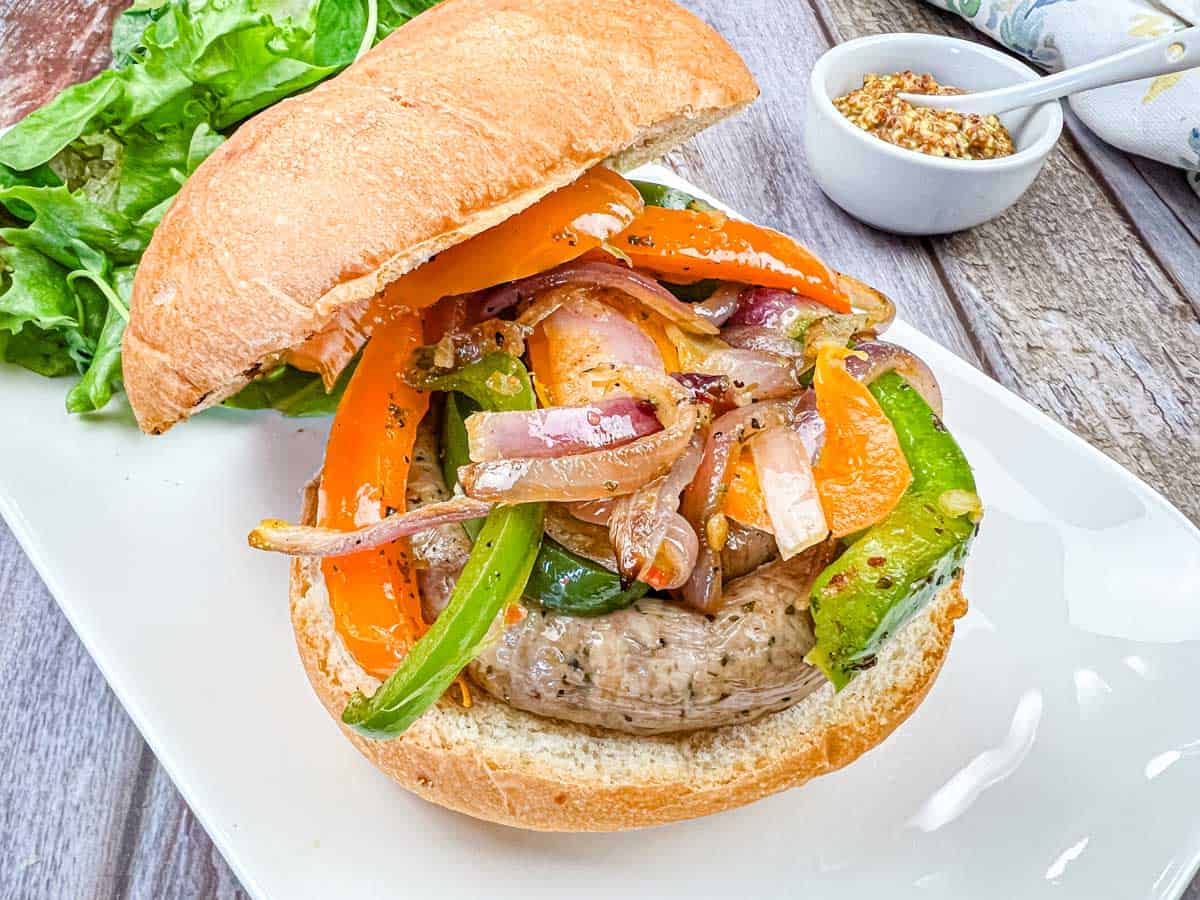 Sausage sandwich topped with grilled bell peppers and onions on a bun, served on a white plate with fresh lettuce and a small bowl of mustard.