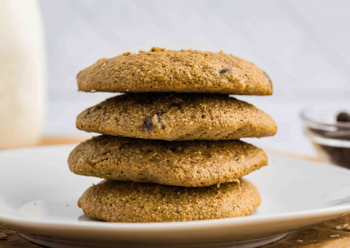 Four chocolate chip cookies are stacked on a white plate, with a blurred bowl and glass in the background.