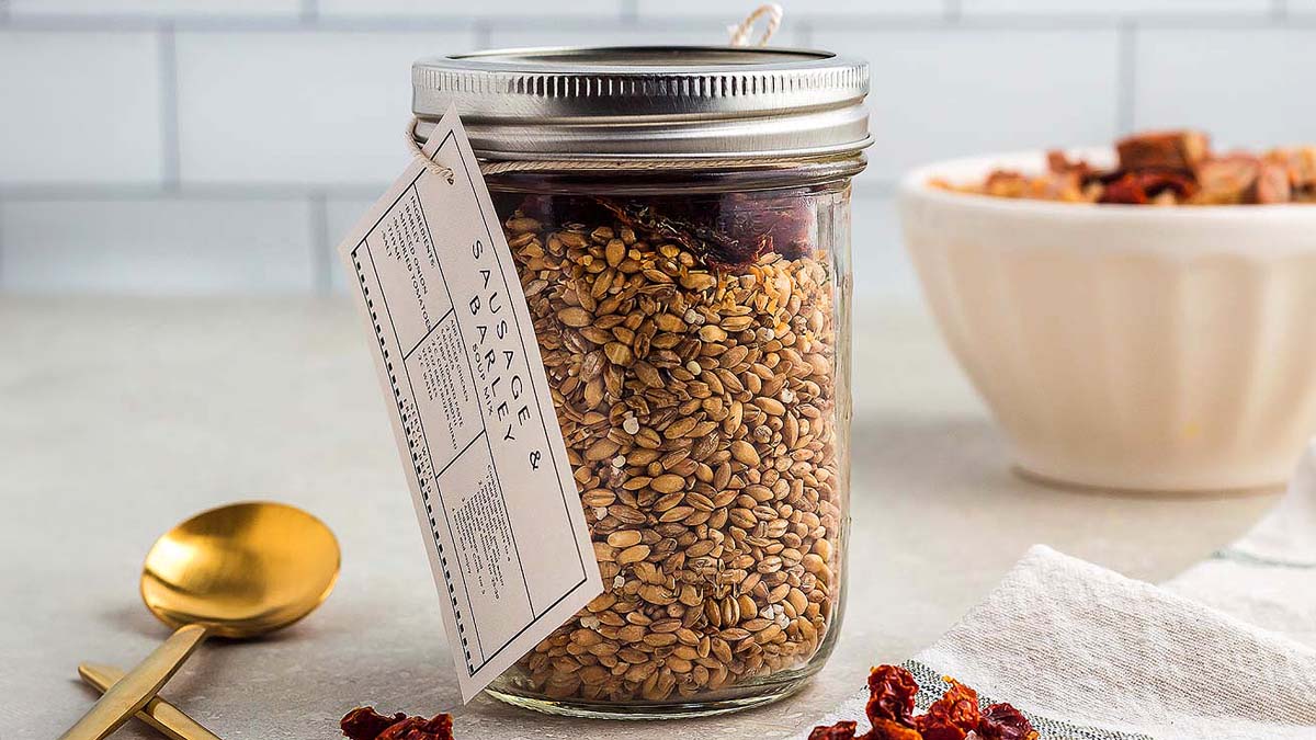 A glass mason jar filled with grains and dried ingredients sits on a countertop, labeled with a tag. A gold spoon and a bowl of similar ingredients are nearby.
