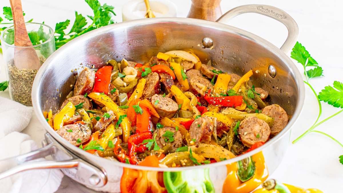 Stainless steel pan filled with cooked sausage slices, red and yellow bell peppers, onions, and herbs on a white countertop with fresh parsley nearby.