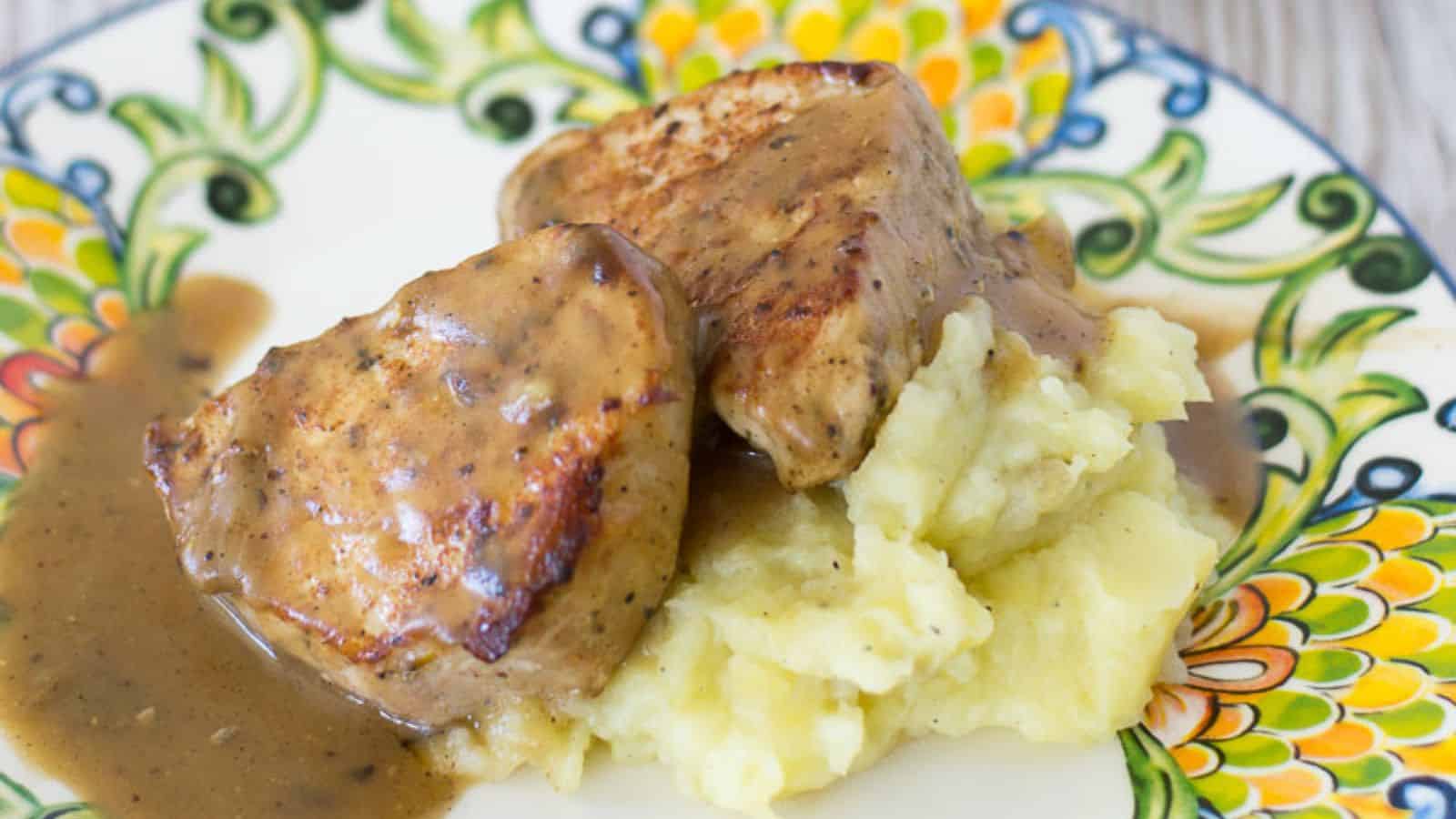 Two pieces of pork with brown gravy served on a mound of mashed potatoes on a decorative plate.
