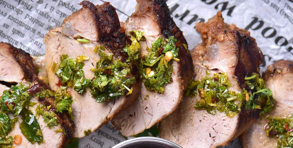 Sliced grilled meat topped with chopped herbs and spices, served on newspaper-style paper, with a metal container of sauce nearby.