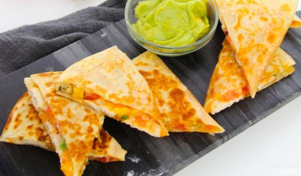 Sliced cheese quesadillas with visible vegetables are arranged on a dark serving board next to a small bowl of guacamole.