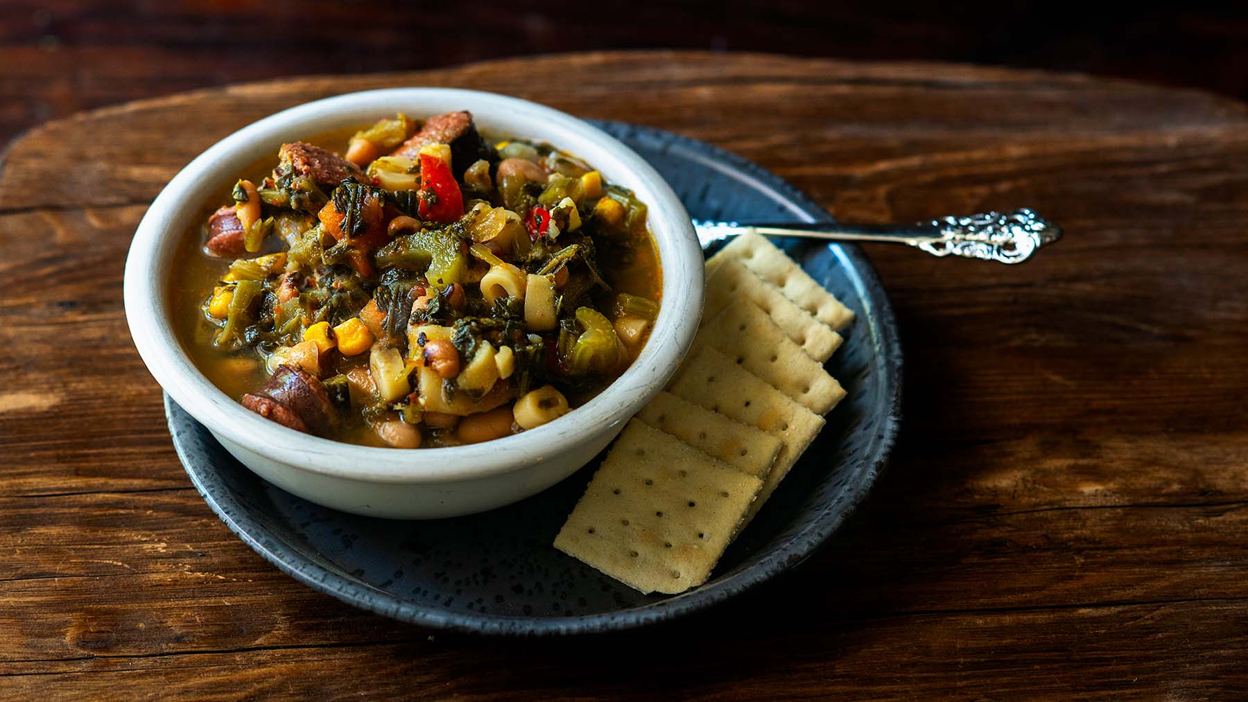 A bowl of vegetable and bean soup with elbow macaroni sits on a plate beside several saltine crackers and a spoon, placed on a wooden table.