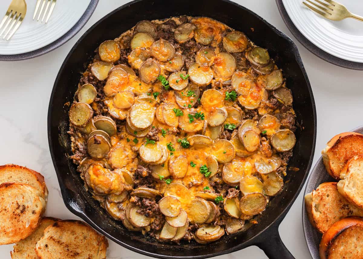 A skillet filled with a baked dish of sliced potatoes, ground beef, and melted cheese, garnished with chopped parsley, surrounded by toasted bread slices.