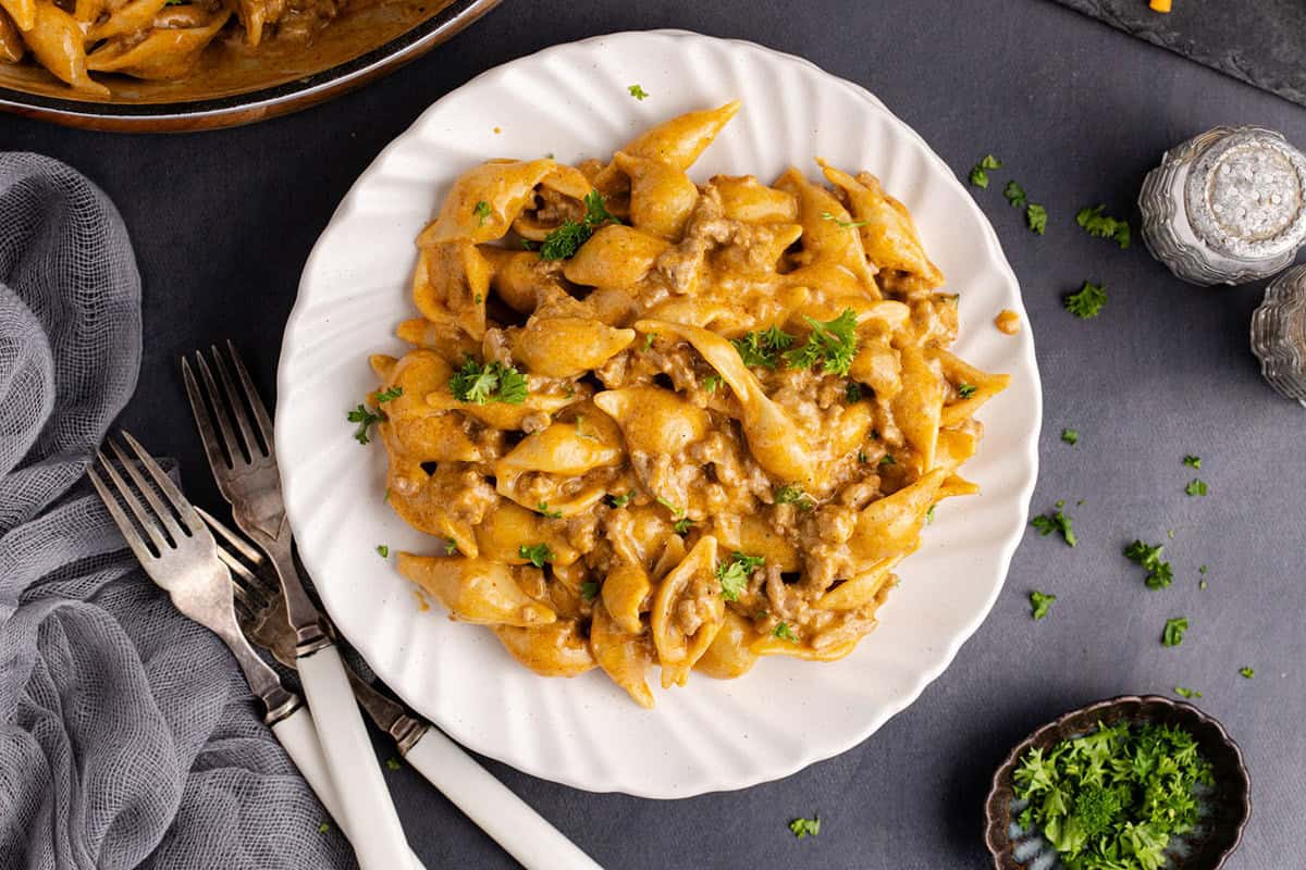 A white plate with creamy pasta shells mixed with ground meat and garnished with chopped parsley, set on a dark surface with cutlery beside it.