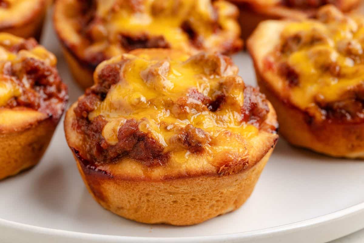 A close-up of baked muffin-shaped cups filled with ground beef and melted cheddar cheese on a white plate.