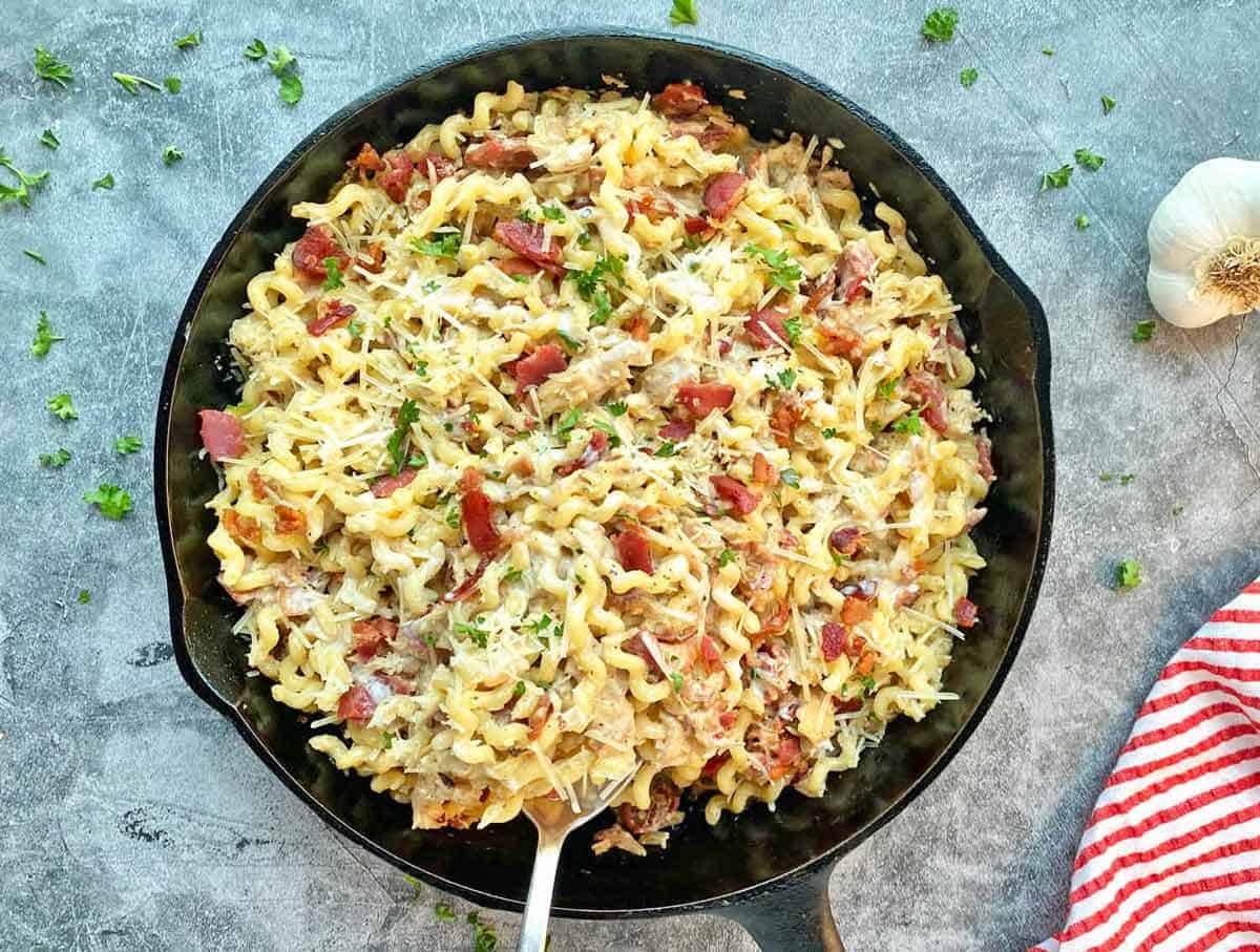 A skillet filled with cooked pasta mixed with bacon, grated cheese, and herbs, with a fork resting inside. Garlic and a red-striped cloth are nearby.