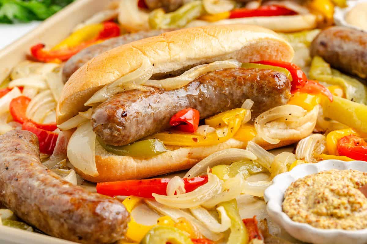 Grilled sausages in buns topped with sautรฉed onions and bell peppers, surrounded by extra sausages and vegetables, with a small dish of mustard on the side.
