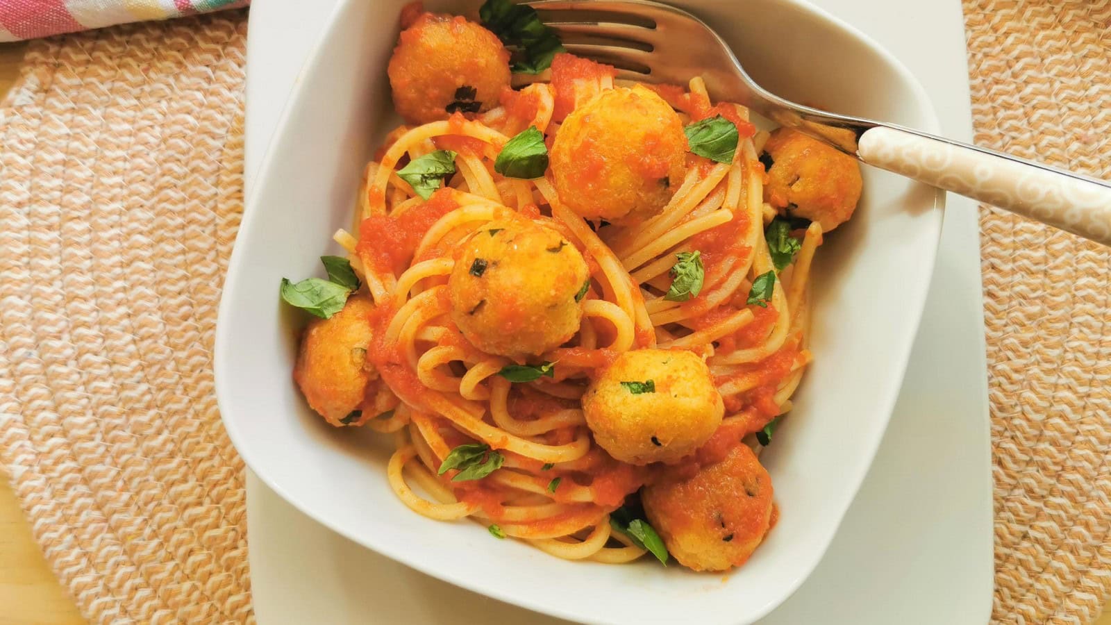 A white bowl of spaghetti topped with tomato sauce, small breaded balls, and chopped basil, with a fork resting on the bowl.