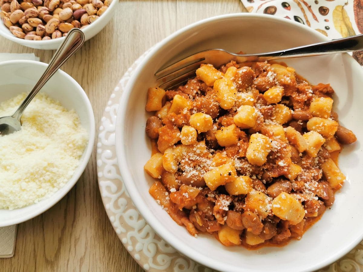 A bowl of gnocchi with tomato sauce and beans, topped with grated cheese, sits on a plate next to a bowl of grated cheese and a bowl of nuts.