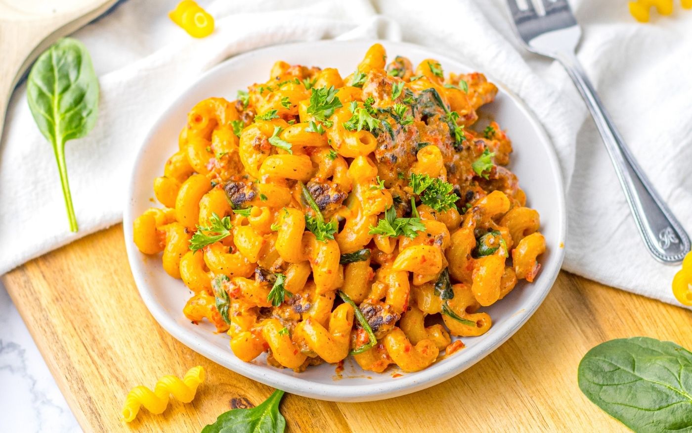 A plate of cooked pasta with a creamy orange sauce, garnished with chopped herbs, sits on a wooden board alongside scattered spinach leaves and a fork.