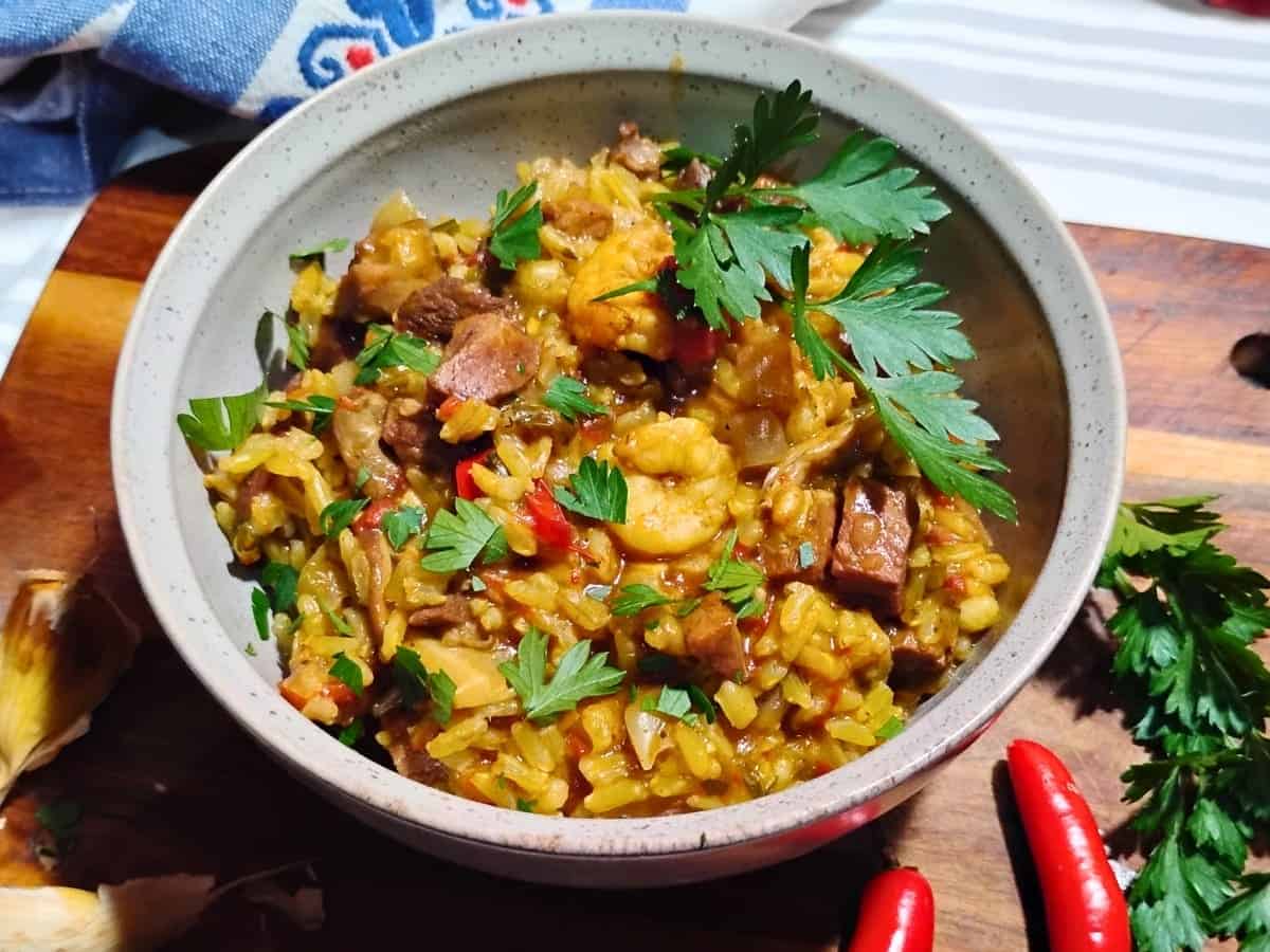 A bowl of jambalaya with rice, shrimp, sausage, vegetables, and fresh parsley, placed on a wooden board beside red chili peppers and garlic.
