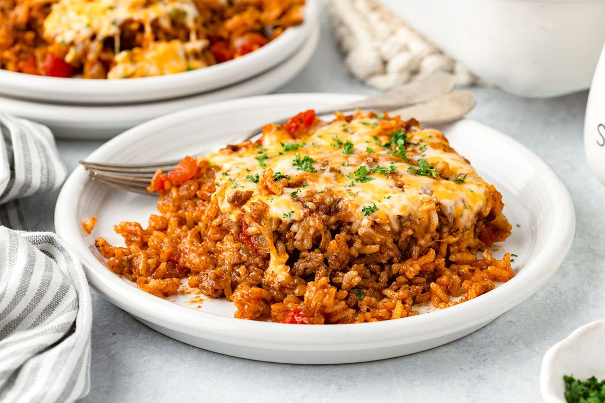 A serving of cheesy ground beef and rice casserole garnished with parsley is on a white plate with a fork.
