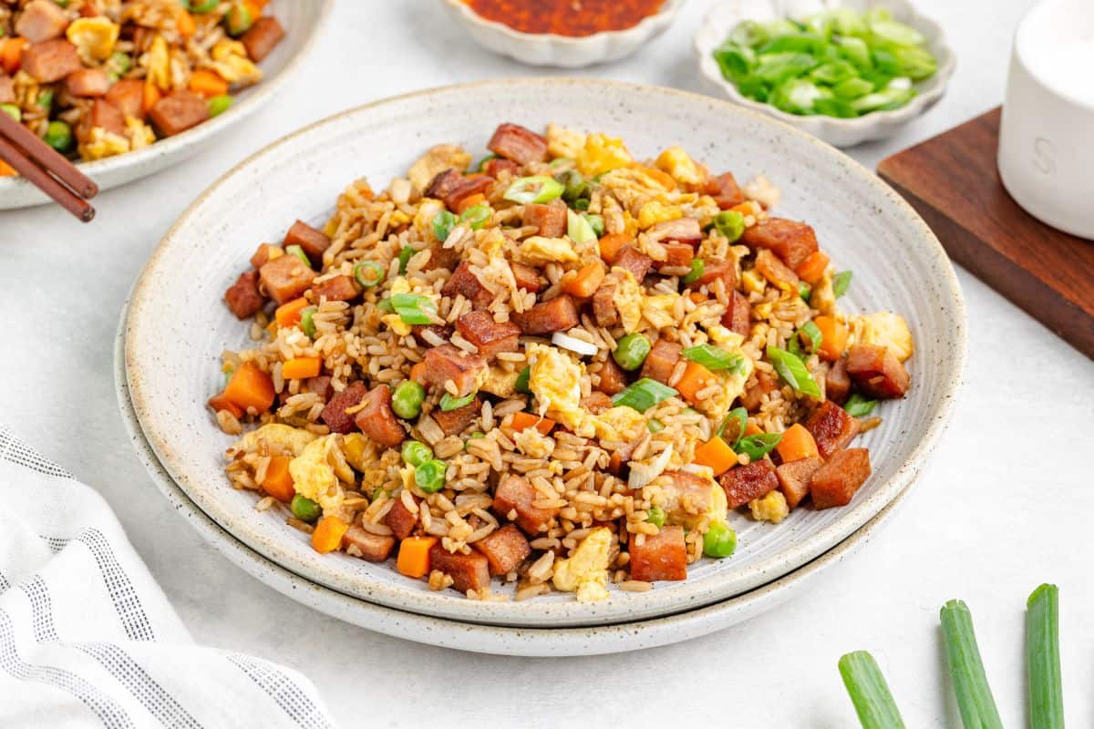 A plate of fried rice with diced ham, scrambled eggs, peas, carrots, and green onions, served on a ceramic dish.
