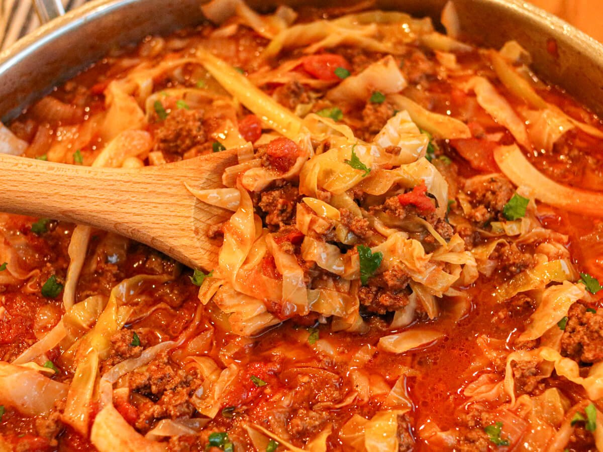 A wooden spoon scoops a mixture of cooked cabbage, ground meat, and tomato sauce from a pot.