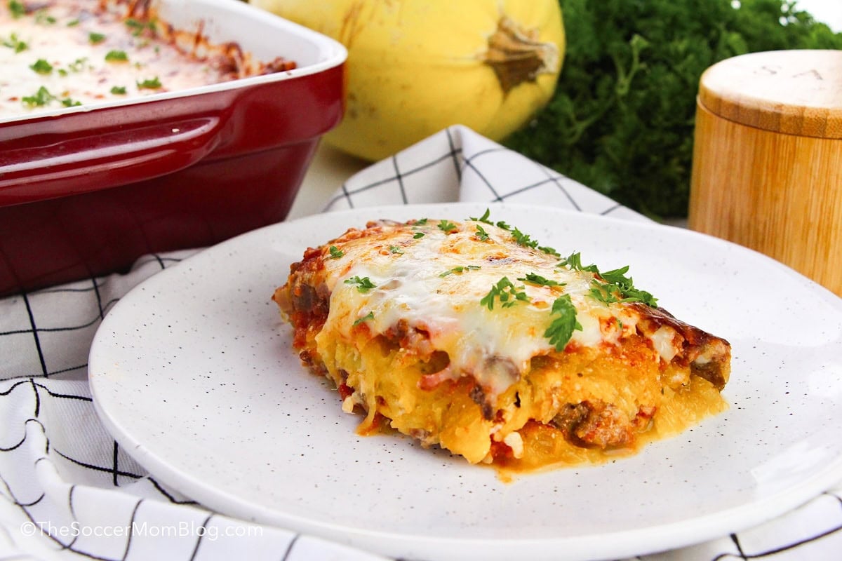 A slice of baked lasagna topped with melted cheese and parsley is served on a white plate, with a casserole dish and squash in the background.
