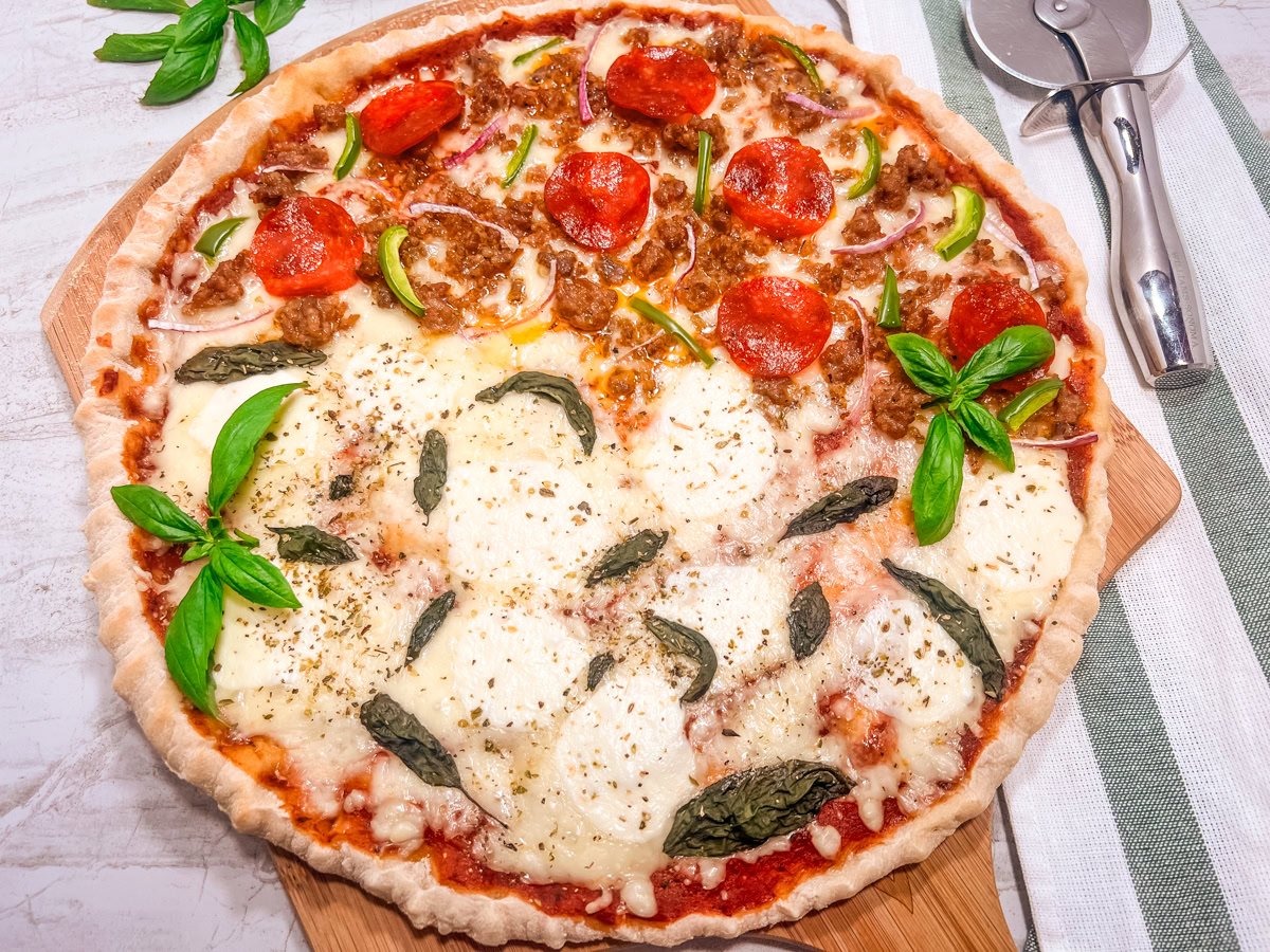 A half-and-half pizza on a wooden board: one side with mozzarella and basil, the other with pepperoni, sausage, onion, and green pepper. A pizza cutter is beside the pizza.