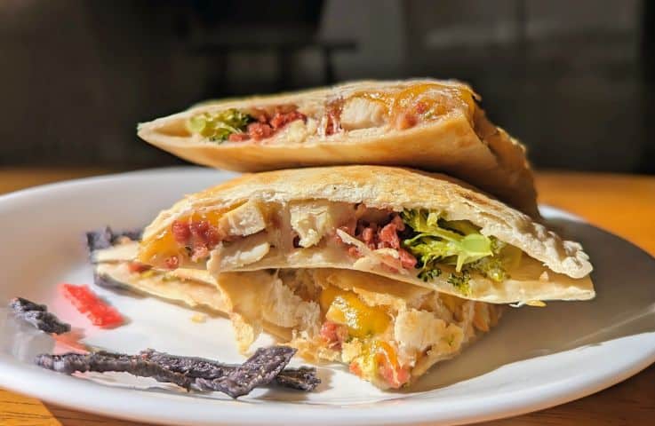 Two pita halves filled with chicken, lettuce, cheese, and bacon are stacked on a white plate, with a few vegetable garnishes on the side.