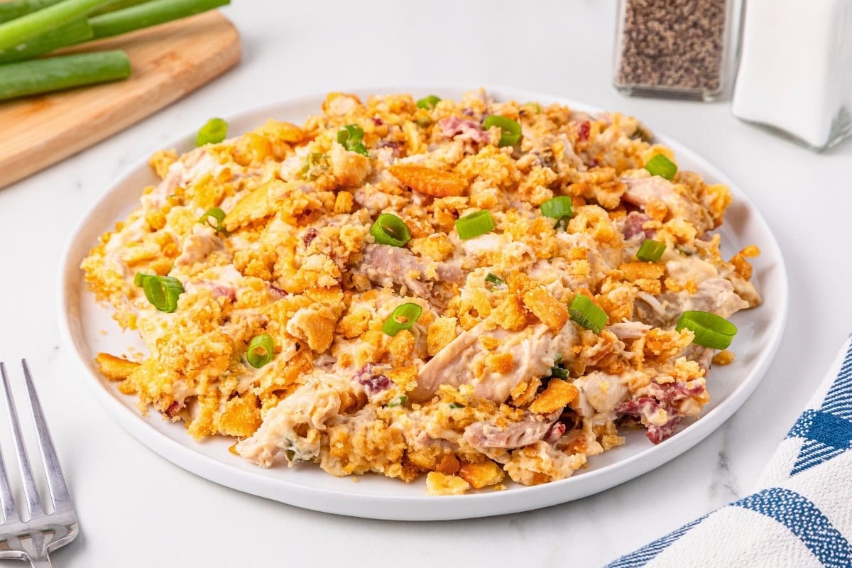A plate of chicken casserole topped with crushed crackers and garnished with chopped green onions sits on a white table next to a fork, salt, pepper, and green onions.