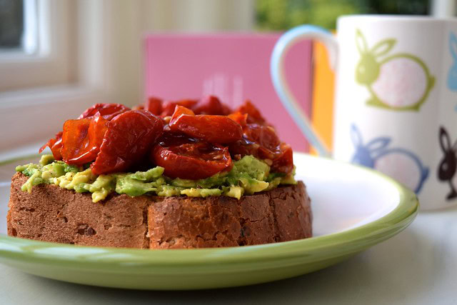 A slice of bread topped with mashed avocado and roasted tomatoes on a green-rimmed plate, next to a mug decorated with colorful bunny illustrations.