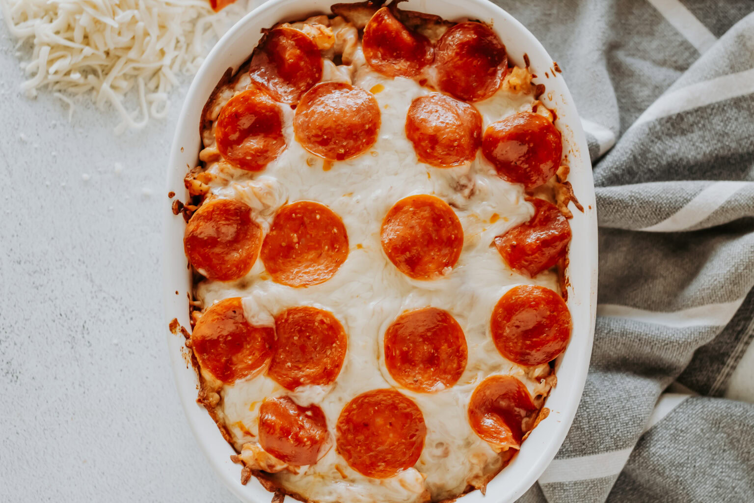 A baked casserole topped with melted cheese and slices of pepperoni in a white oval dish, next to a gray-striped towel and shredded cheese.